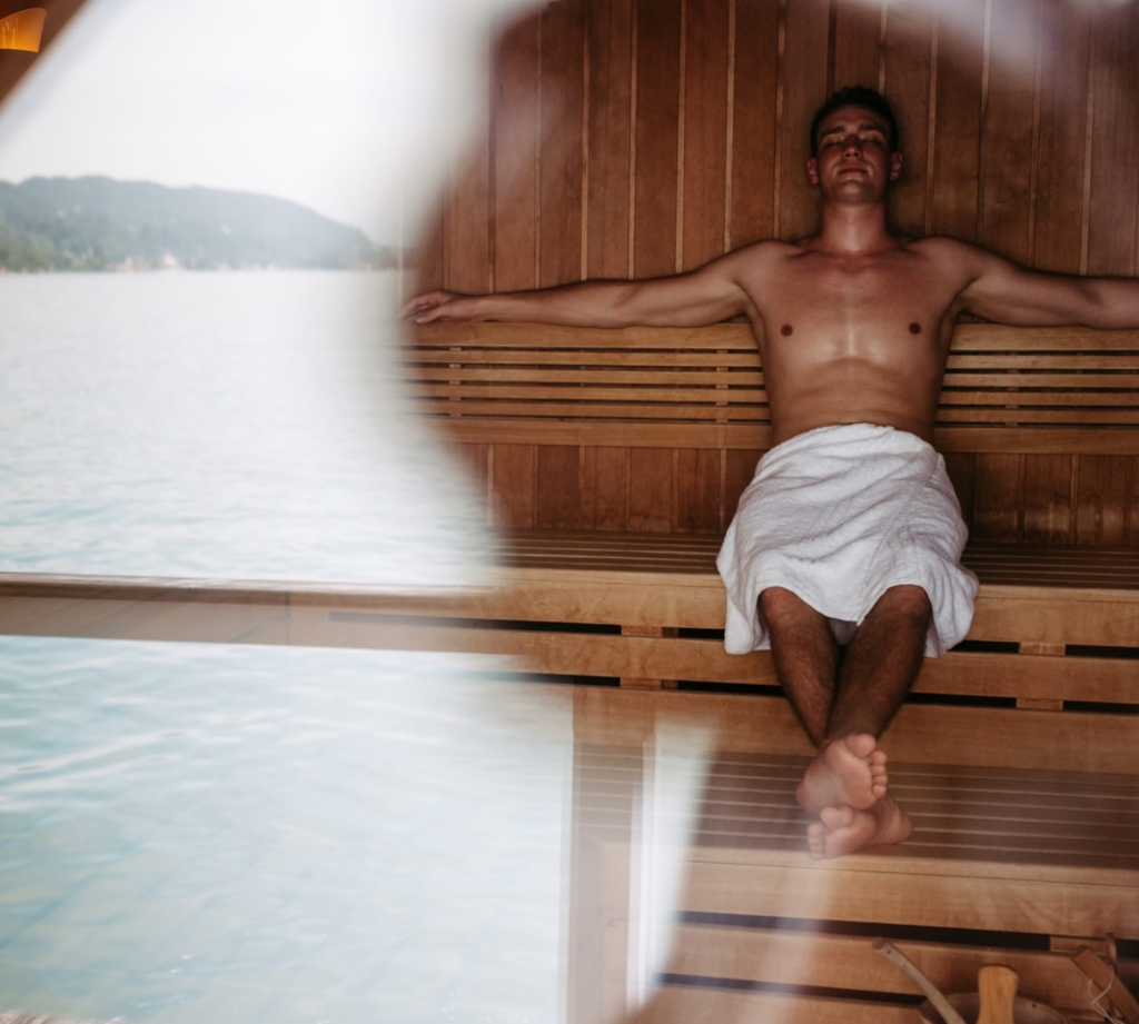 Mann sitzt in einer Seesauna und im Glas spiegelt sich der Wörthersee wieder.