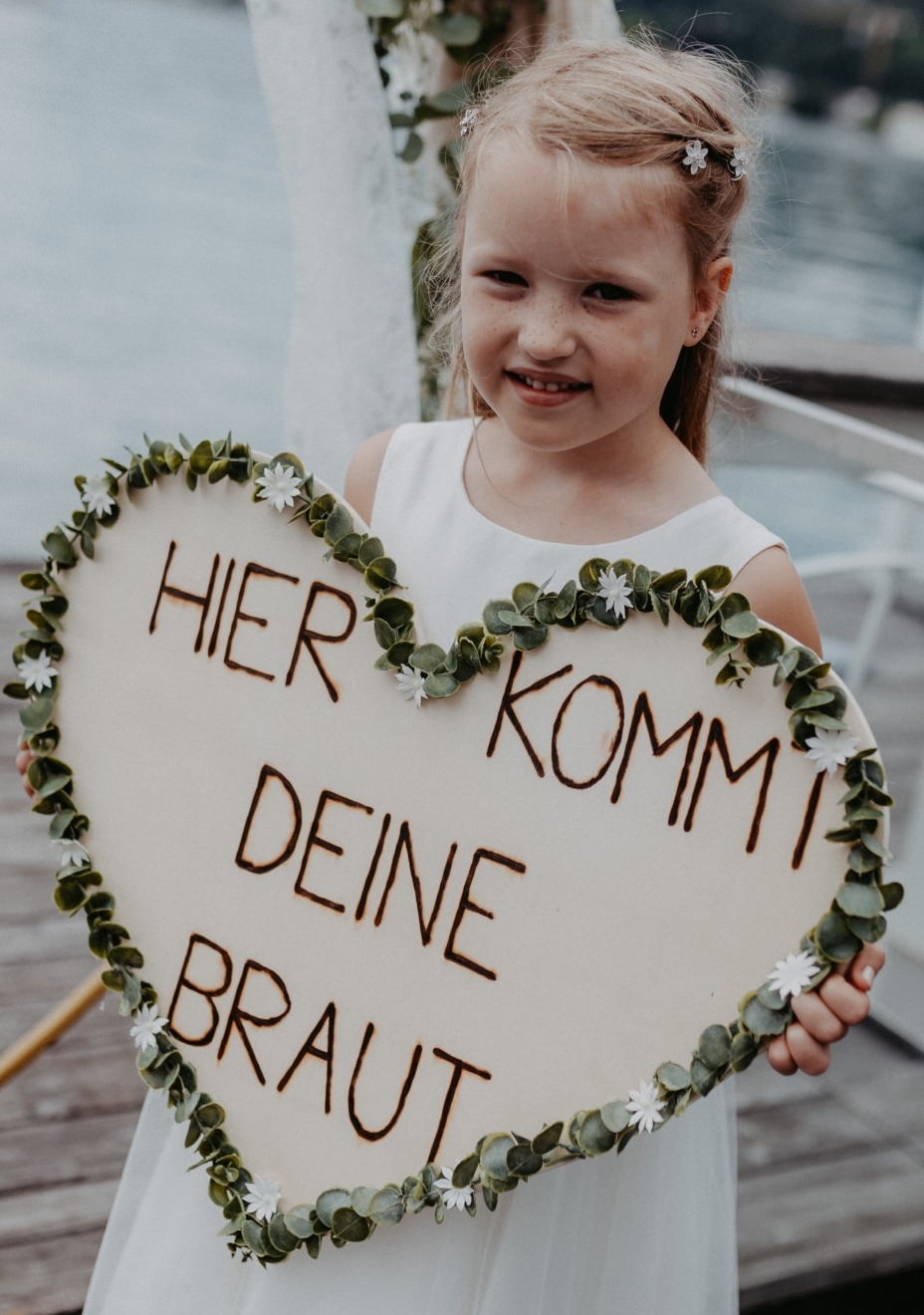 Ein kleines Mädchen trägt ein großes herzförmiges Schild mit der Aufschrift 'Hier kommt deine Braut'.