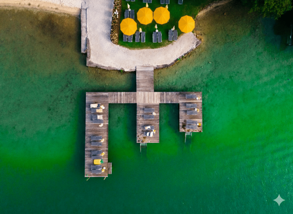 Luftaufnahme eines Holzstegs mit Liegen und Sonnenschirmen über grünem Wasser.