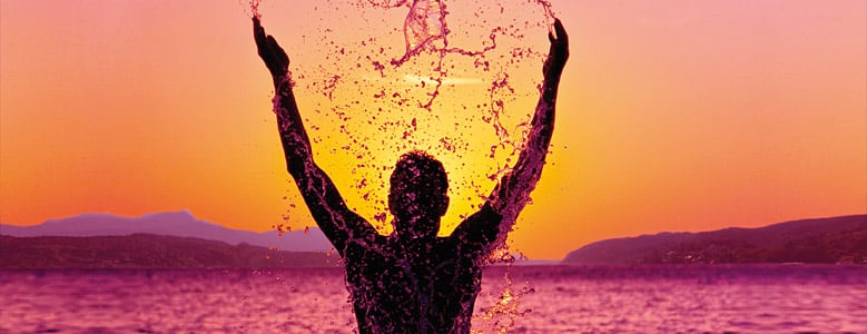 Eine silhouettierte Person, die Wasser über ihren Kopf spritzt, während die Sonne untergeht.